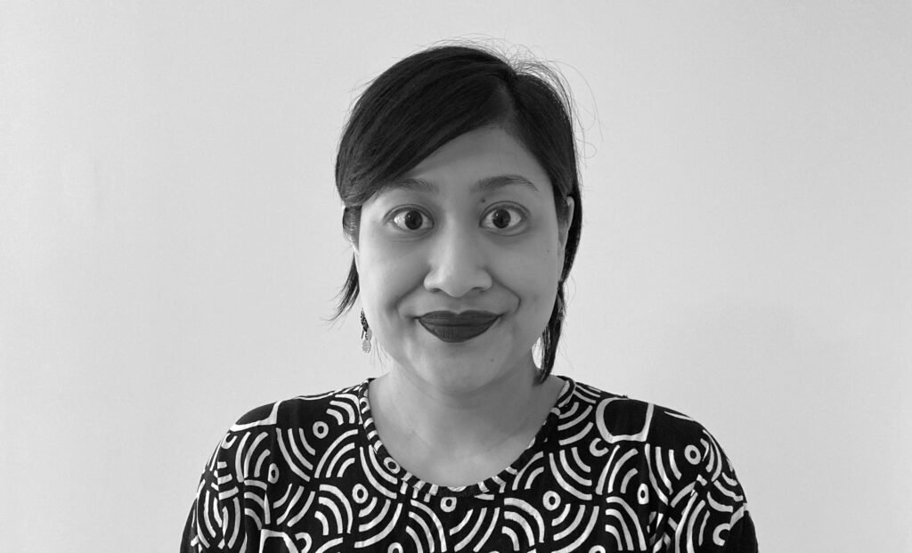 A black and white photo of an Indonesian woman with black hair tied back. She is wearing lipstick, and a black and white daster, and is smiling to the camera against a white background.
