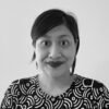 A black and white photo of an Indonesian woman with black hair tied back. She is wearing lipstick, and a black and white daster, and is smiling to the camera against a white background.