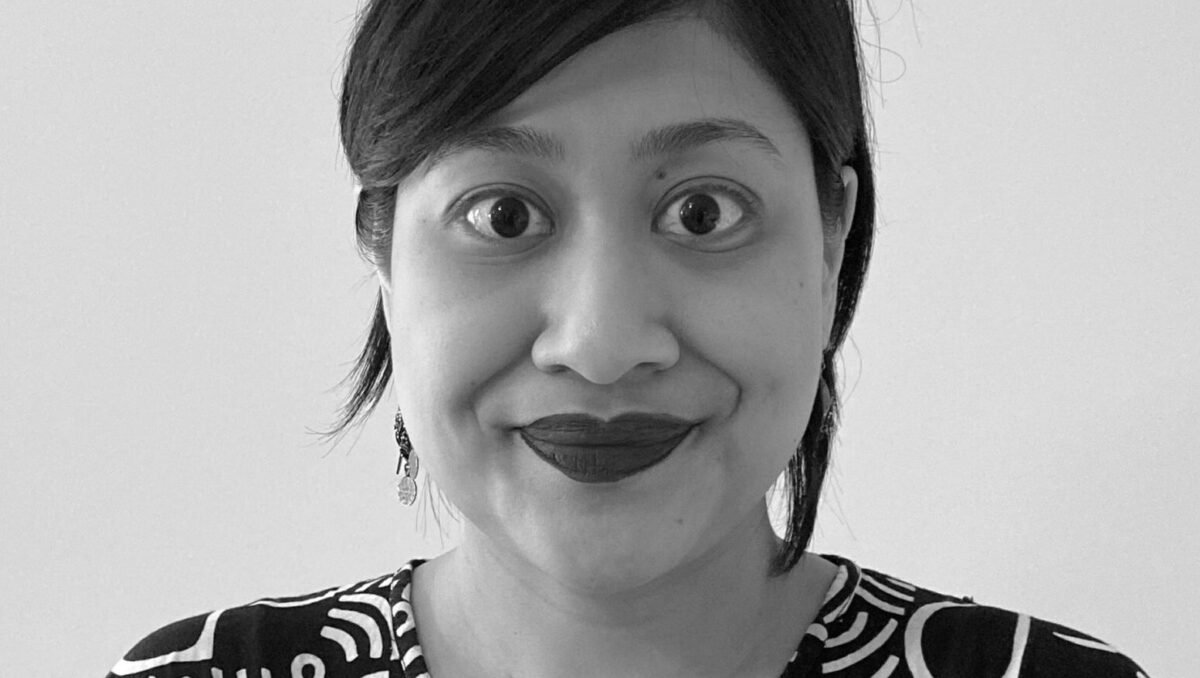 A black and white photo of an Indonesian woman with black hair tied back. She is wearing lipstick, and a black and white daster, and is smiling to the camera against a white background.