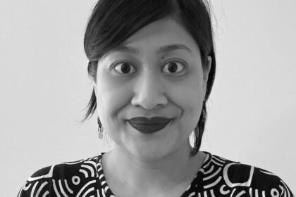 A black and white photo of an Indonesian woman with black hair tied back. She is wearing lipstick, and a black and white daster, and is smiling to the camera against a white background.