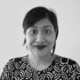 A black and white photo of an Indonesian woman with black hair tied back. She is wearing lipstick, and a black and white daster, and is smiling to the camera against a white background.