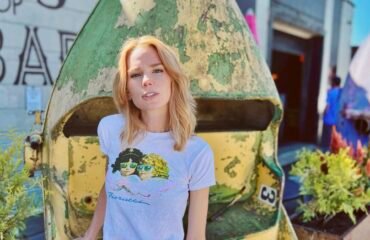 Blonde Caucasian woman (author Claire Hopple), wearing a graphic tee shirt and skirt, stands facing the camera. Her right hand rests on a vertically turned metallic cart behind her, with planters on either side.