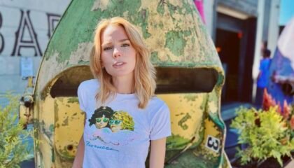 Blonde Caucasian woman (author Claire Hopple), wearing a graphic tee shirt and skirt, stands facing the camera. Her right hand rests on a vertically turned metallic cart behind her, with planters on either side.