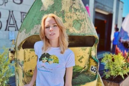 Blonde Caucasian woman (author Claire Hopple), wearing a graphic tee shirt and skirt, stands facing the camera. Her right hand rests on a vertically turned metallic cart behind her, with planters on either side.