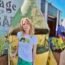 Blonde Caucasian woman (author Claire Hopple), wearing a graphic tee shirt and skirt, stands facing the camera. Her right hand rests on a vertically turned metallic cart behind her, with planters on either side.