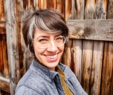 Brunette woman (Stefanie Kirby) with short hair and a blue shirt grins at camera.