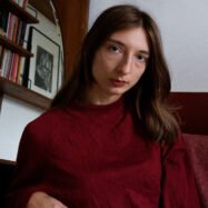 Brunette woman (Nasim Luczaj) in red top looks at camera.