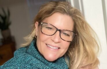 Blonde woman in blue turtleneck smiles into the camera.