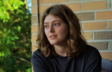 Woman in a black t shirt looks off camera, in front of a brick wall.