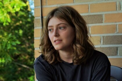 Woman in a black t shirt looks off camera, in front of a brick wall.