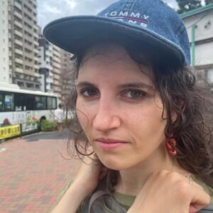 Woman with curly brown hair wearing a baseball cap looks at the camera on a city street.