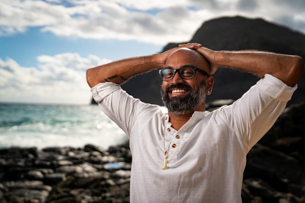 Man stands at seashore in a white shirt with a big grin, resting his hands on his head.