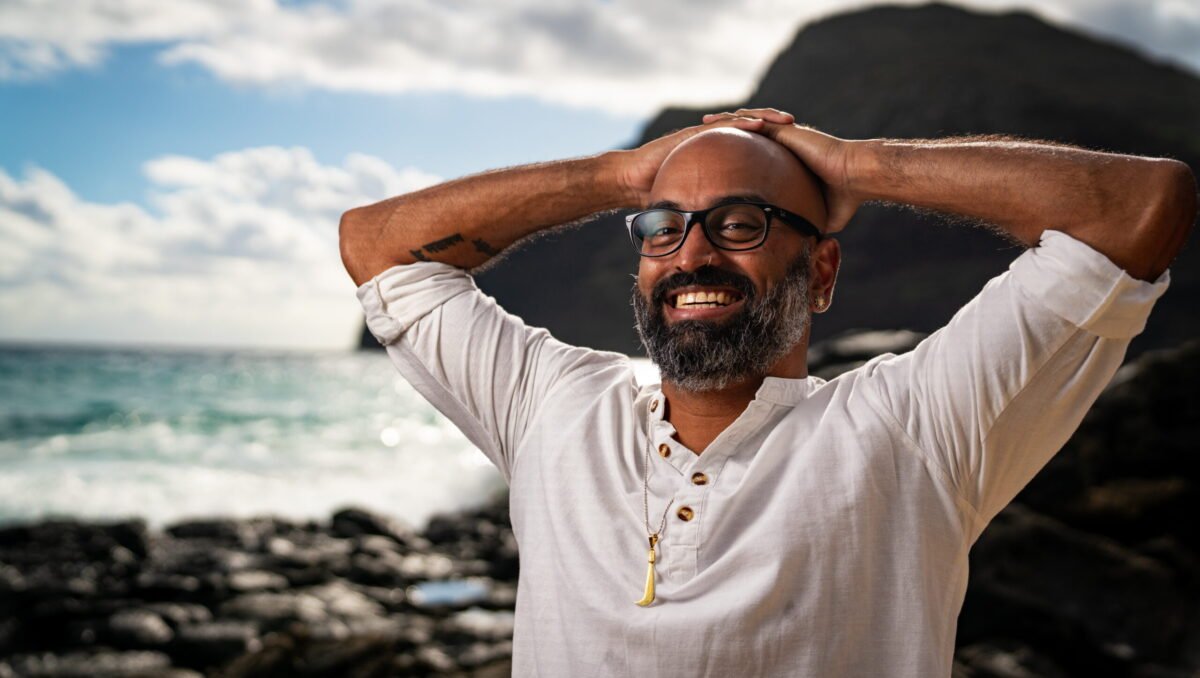 Man stands at seashore in a white shirt with a big grin, resting his hands on his head.