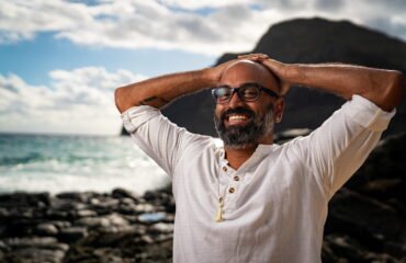 Man stands at seashore in a white shirt with a big grin, resting his hands on his head.