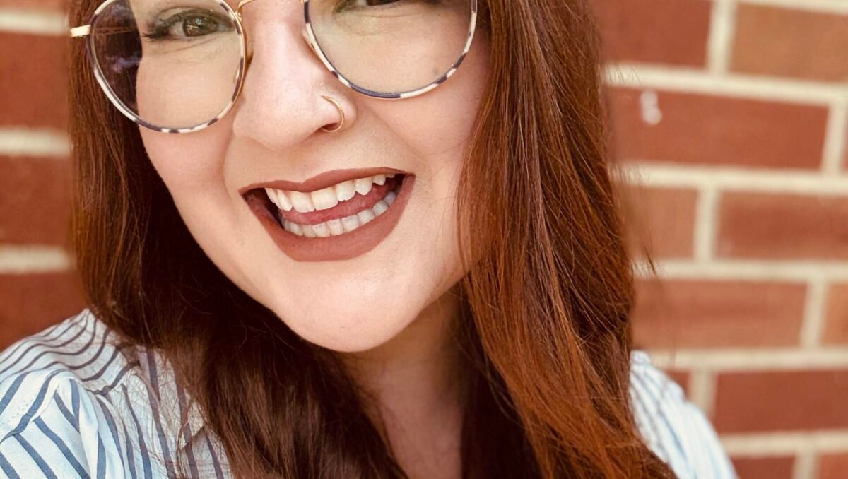 Red haired woman smiles widely in front of a brick wall.