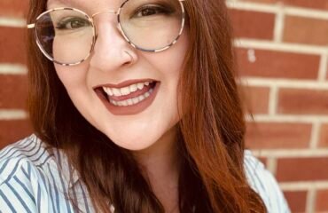 Red haired woman smiles widely in front of a brick wall.