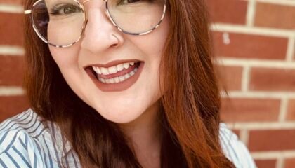 Red haired woman smiles widely in front of a brick wall.