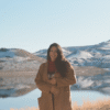 Woman in tan jacket stands in front of mountains and lake.