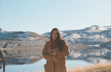 Woman in tan jacket stands in front of mountains and lake.