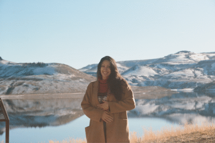 Woman in tan jacket stands in front of mountains and lake.