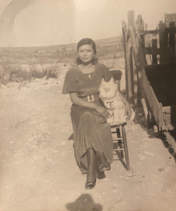 An old, sepia-tone photograph of a young woman seated on a wooden chair by a wooden cart, with a ceramic dog under her arm and the desert in the background.