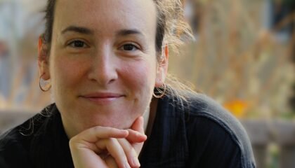 White woman stares directly in the camera, chin on her hand.