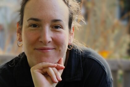 White woman stares directly in the camera, chin on her hand.