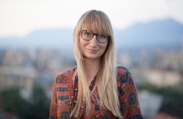 Woman with glasses and bangs looks at the camera, smiling.