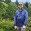 White man in blue jacket and baseball cap stands in front of a grove of trees.
