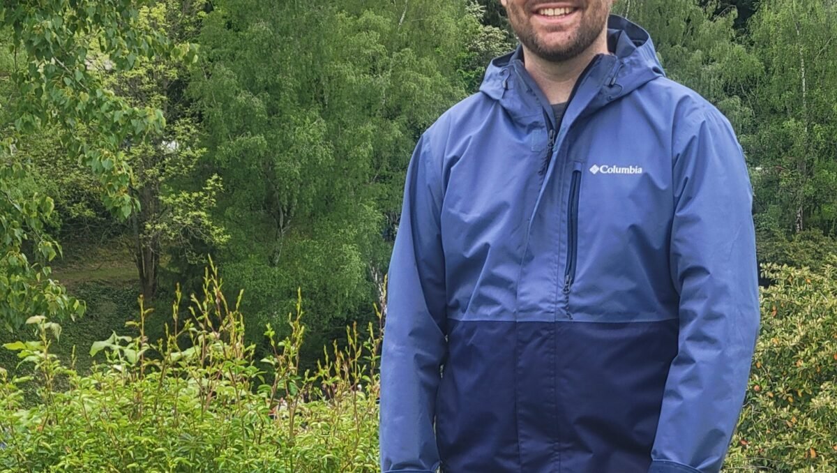 ryan_bender-murphy White man in blue jacket and baseball cap stands in front of a grove of trees.