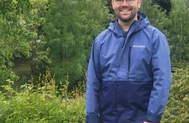 White man in blue jacket and baseball cap stands in front of a grove of trees.