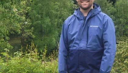 White man in blue jacket and baseball cap stands in front of a grove of trees.