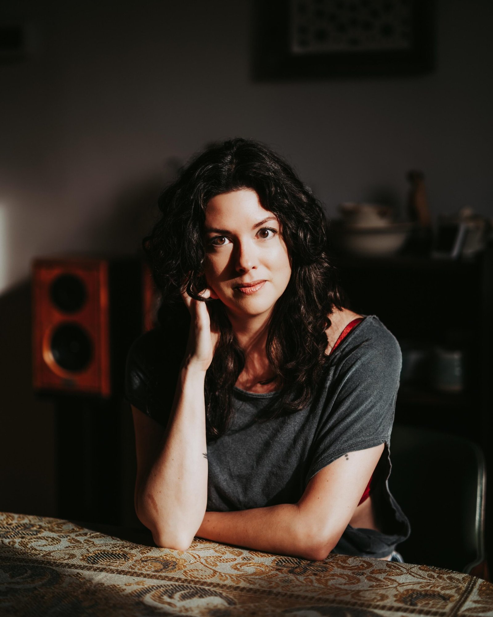Woman with dark hair in a grey t shirt faces the camera, seated at a wooden table.